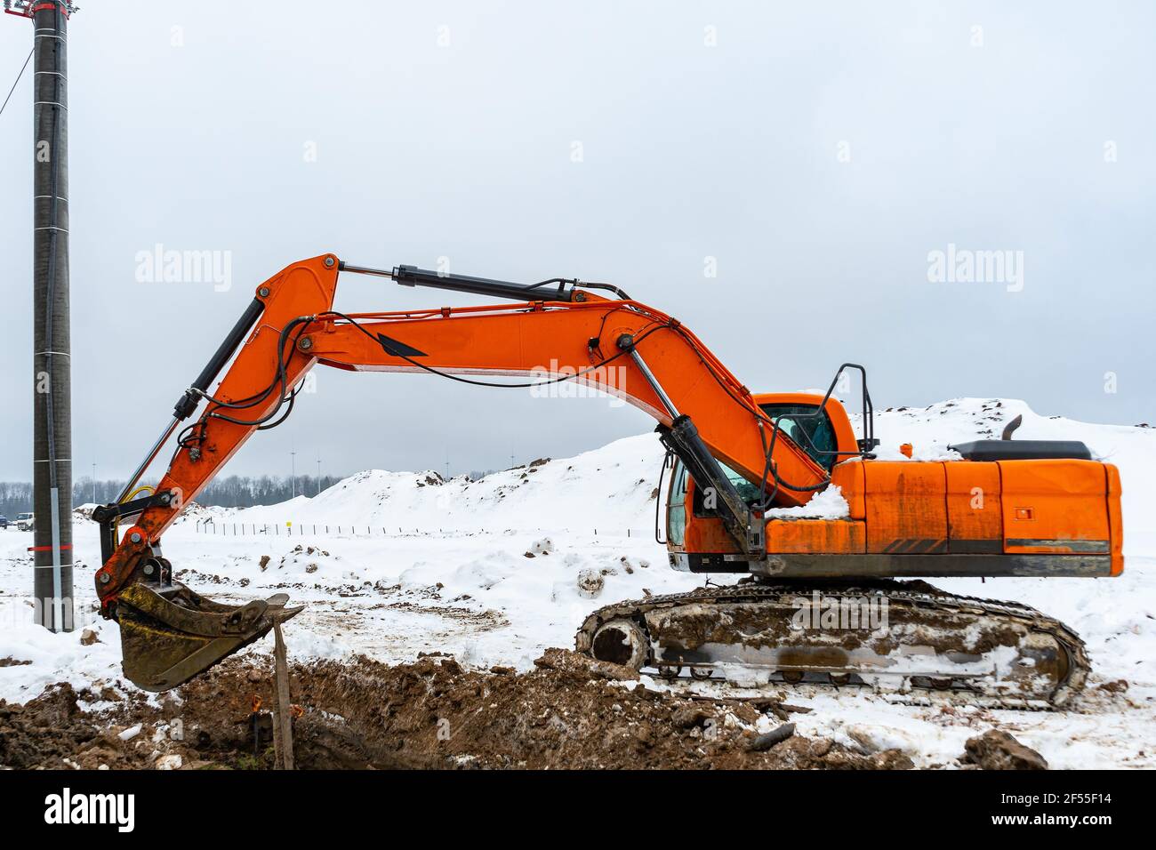 Un grande escavatore arancione scava un buco in un cantiere in inverno. Digger meccanico arancione e foro Foto Stock