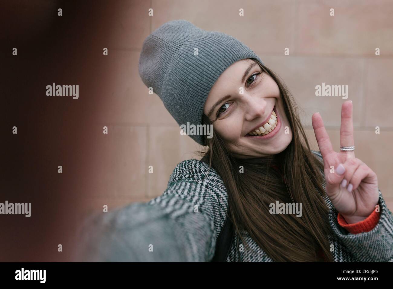 Giovane donna allegra in abiti caldi che mostra segno di pace contro parete Foto Stock