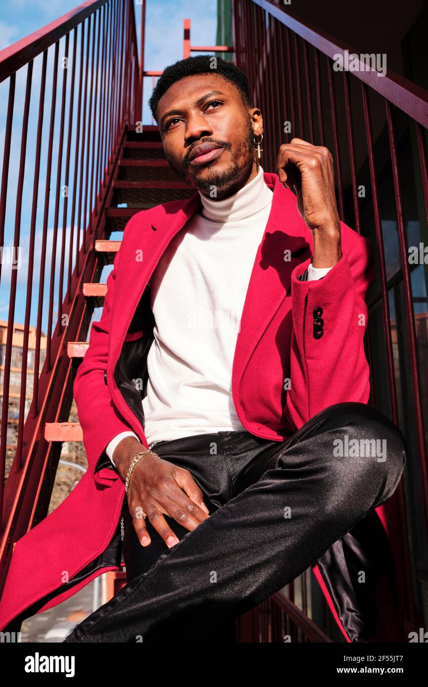 Uomo africano pensieroso con trench rosso seduto su metallo scala durante il giorno di sole Foto Stock