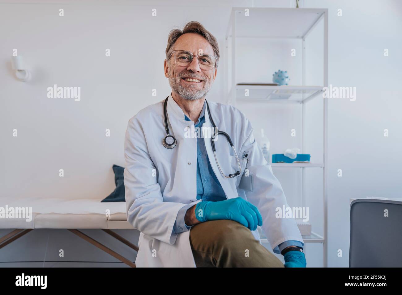 Medico generico con guanto protettivo sorridente mentre si siede in clinica Foto Stock