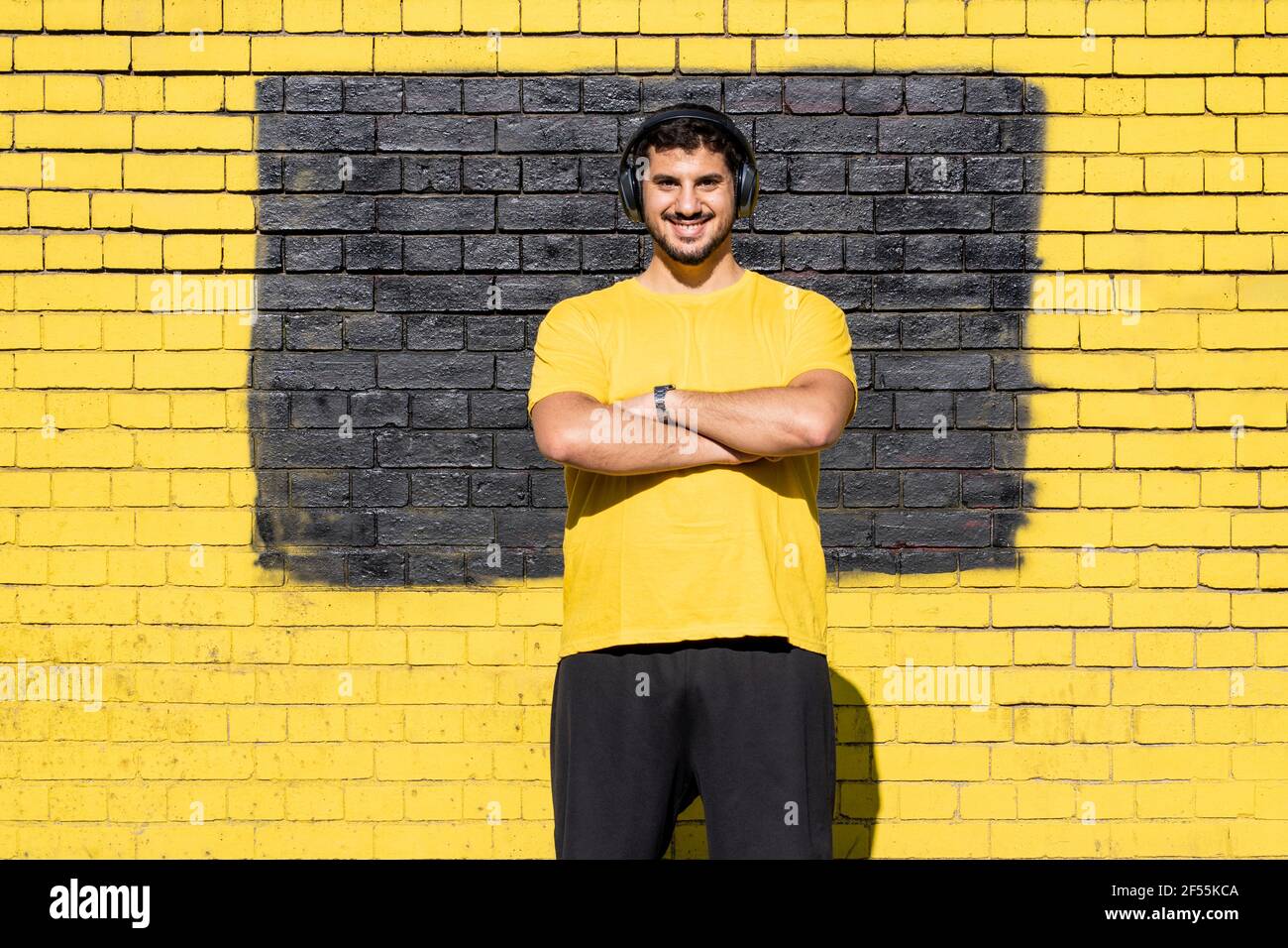 Uomo sorridente con cuffie in piedi contro parete di mattoni gialli Foto Stock