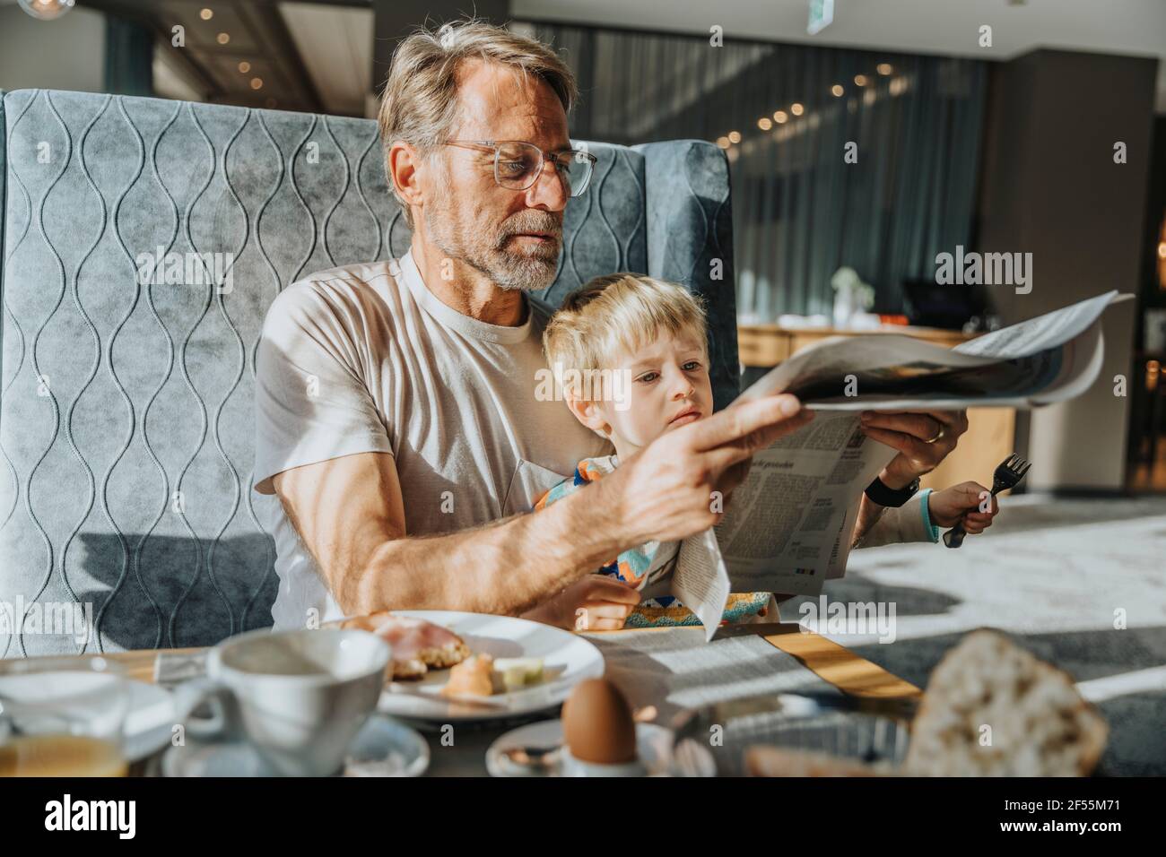 Uomo maturo con il figlio che legge giornale mentre si siede a tavola in camera d'albergo Foto Stock