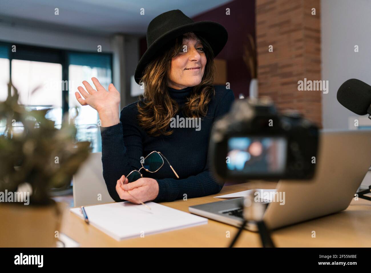 Donna matura che indossa il cappello ondeggiando la mano mentre filma attraverso la macchina fotografica a casa Foto Stock