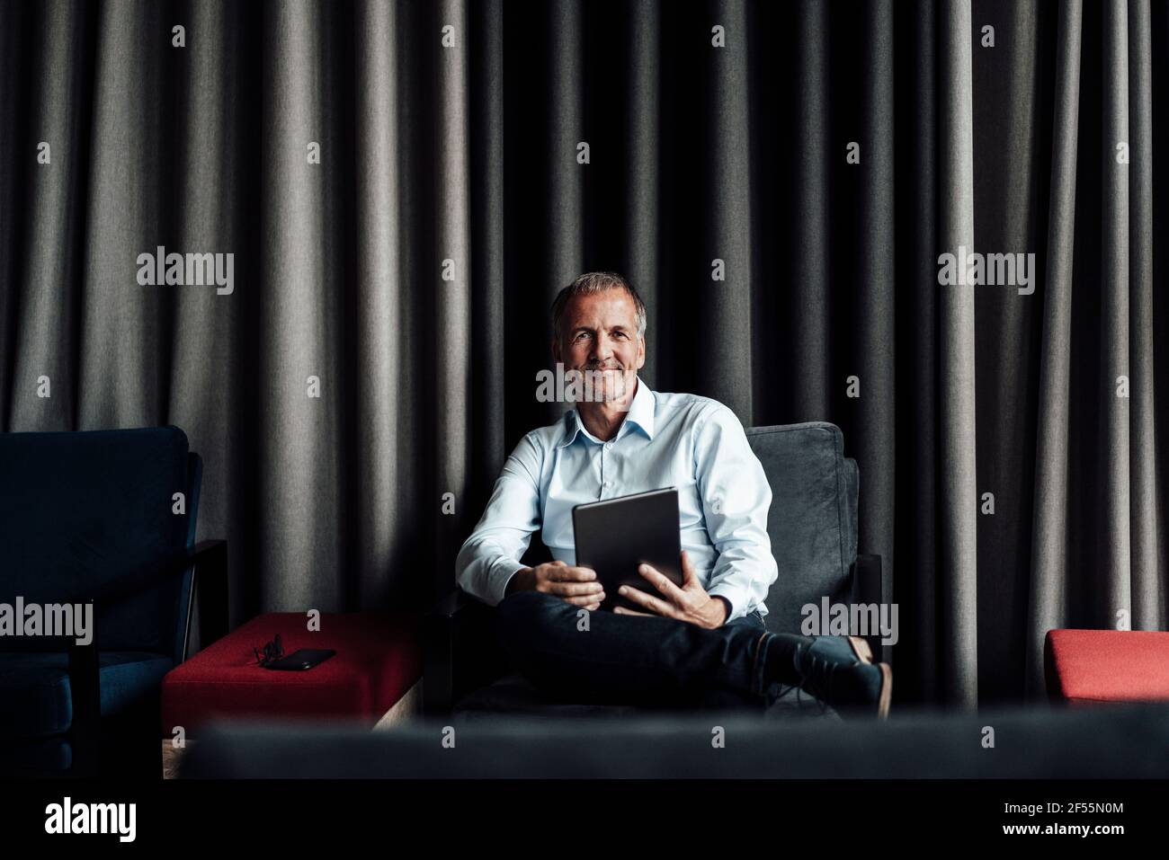 Sorridente professionista senior aziendale che tiene un tablet digitale mentre si è seduti su poltrona in caffetteria d'ufficio Foto Stock
