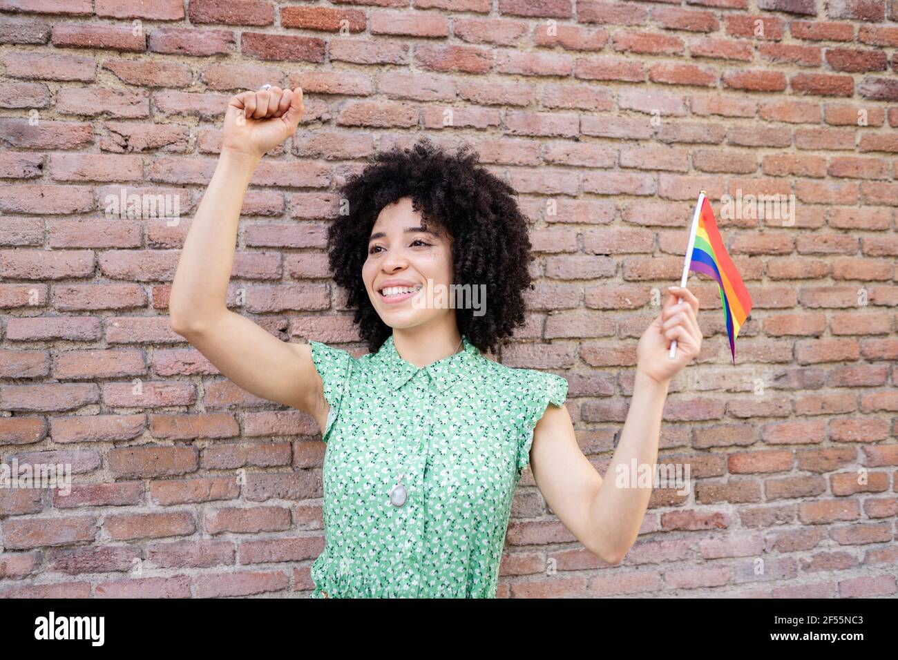 Giovane donna allegra pugno mentre tiene la bandiera iridata LGBTQI contro muro di mattoni Foto Stock