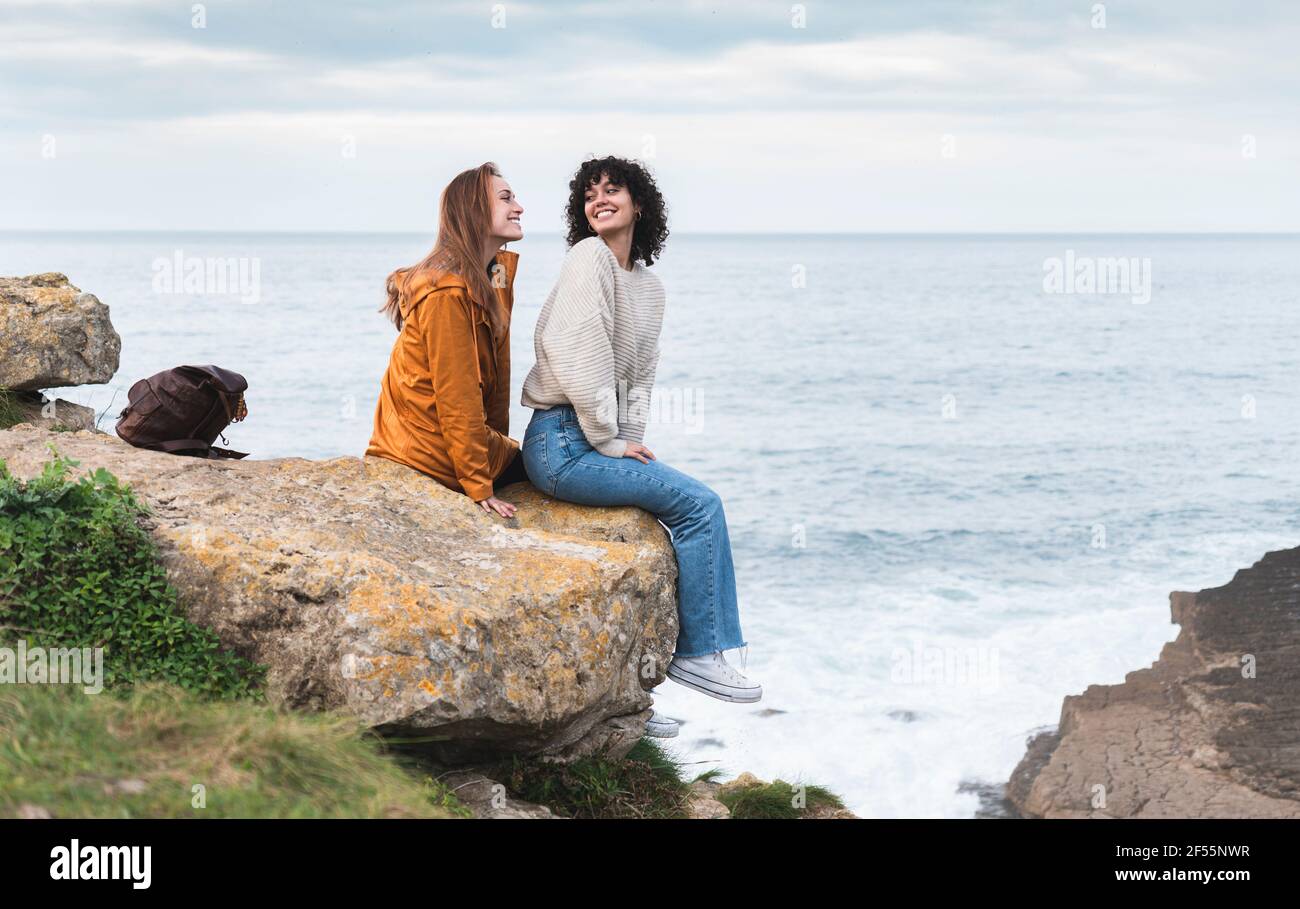 Le amiche sorridono guardandosi mentre si siedono scogliera sul mare Foto Stock