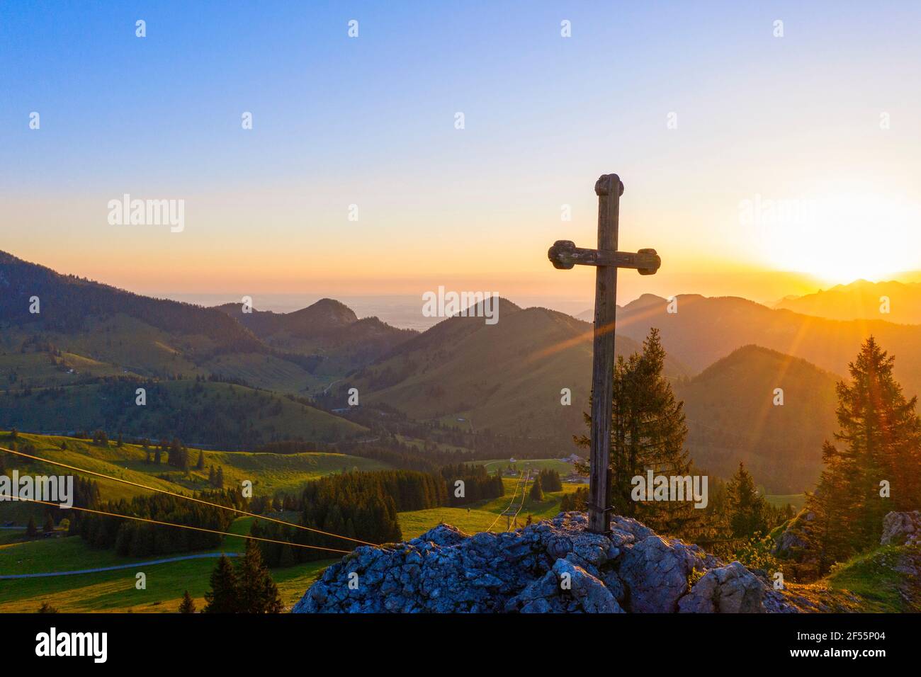 Il sole che sorge illumina la croce cristiana in piedi sulla cima del monte Sudelfeldkopf Foto Stock