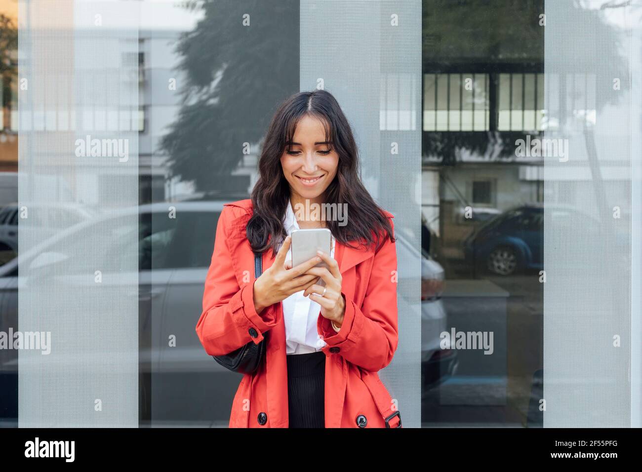 Donna sorridente in trench rosso utilizzando smartphone mentre in piedi contro parete di vetro Foto Stock