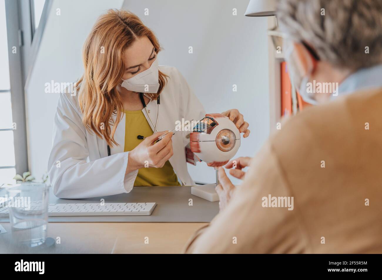 Oftalmologo femminile con maschera facciale che spiega sopra il modello dell'occhio a. paziente seduto in clinica Foto Stock