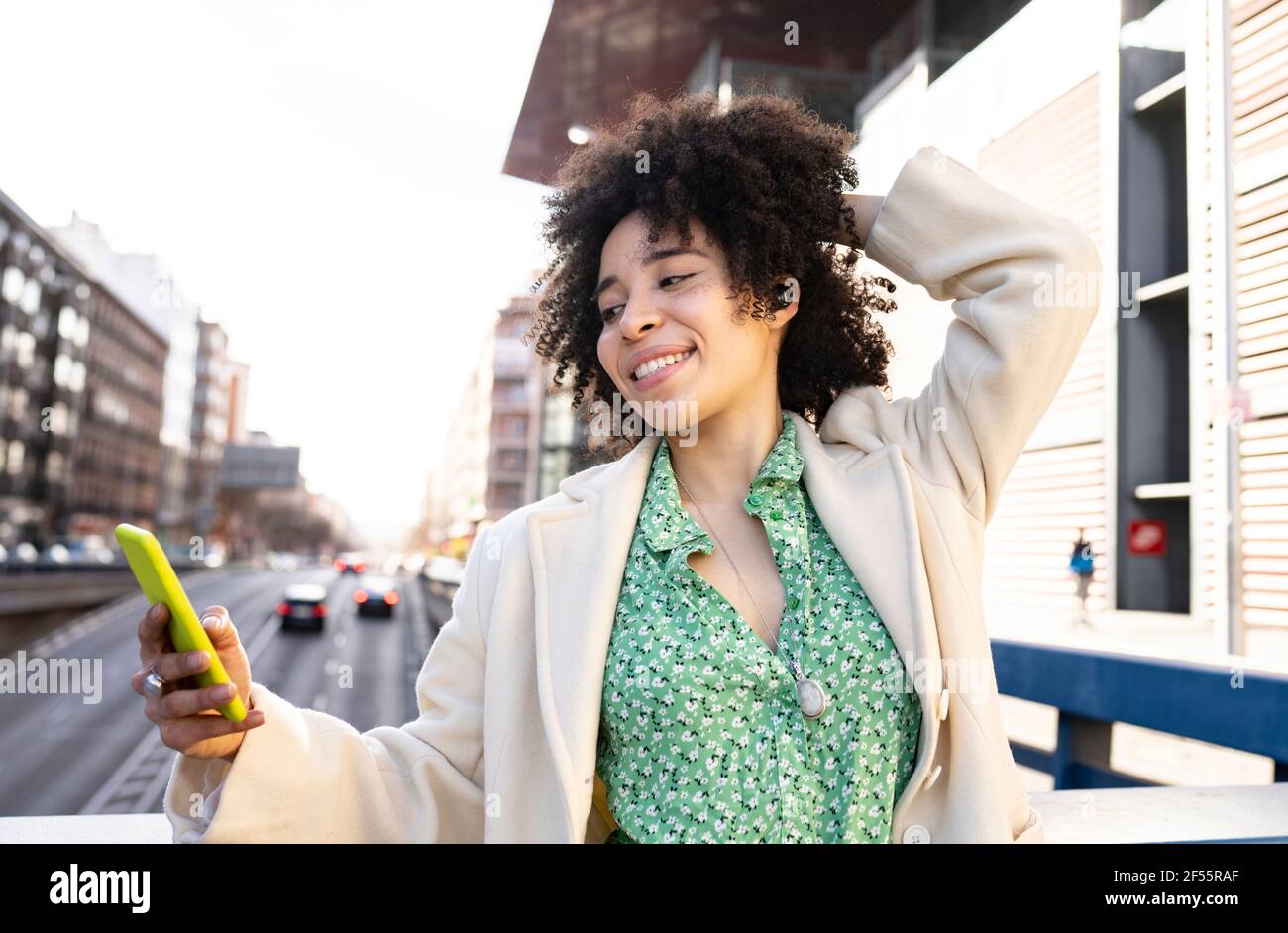Giovane donna con i capelli a mano che ascolta la musica tramite Bluetooth auricolari mentre si utilizza uno smartphone in città Foto Stock