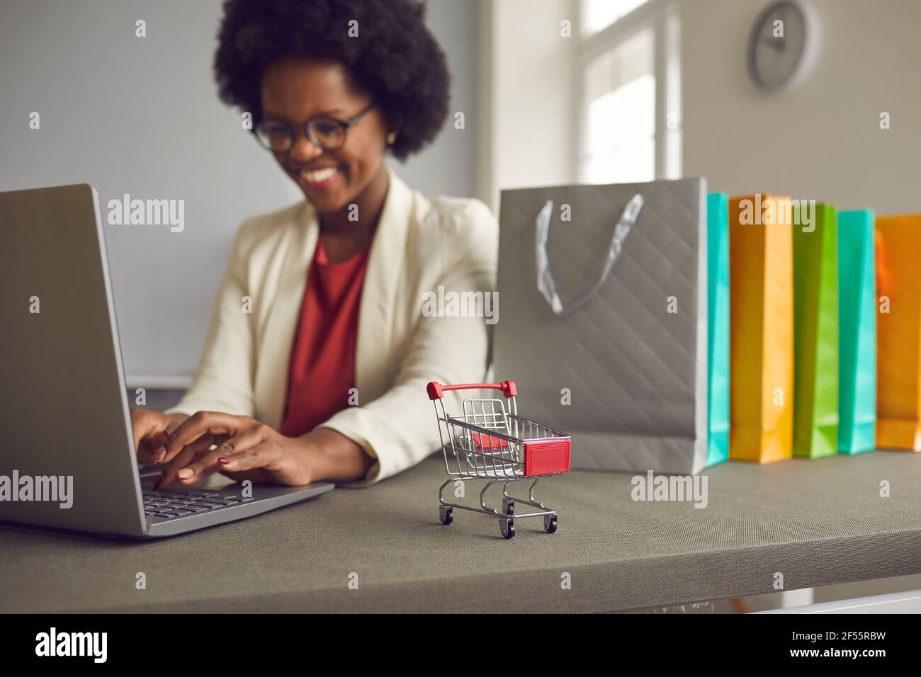 Carrello giocattolo della drogheria in piedi su un tavolo vicino alle borse della spesa e vicino ad una donna che acquista in linea. Foto Stock