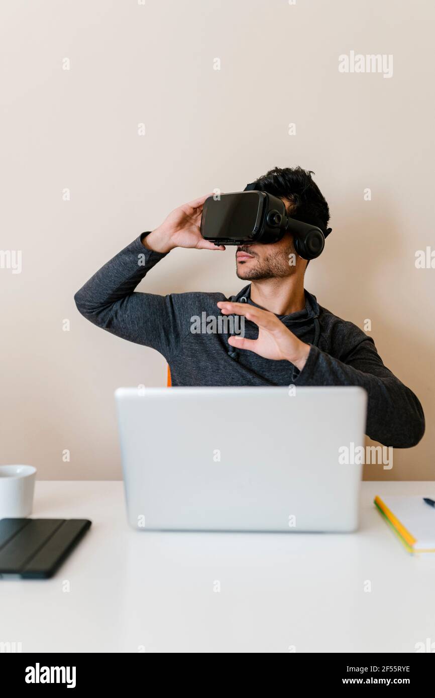 Uomo seduto con un laptop che indossa un simulatore di realtà virtuale al tavolo in soggiorno Foto Stock