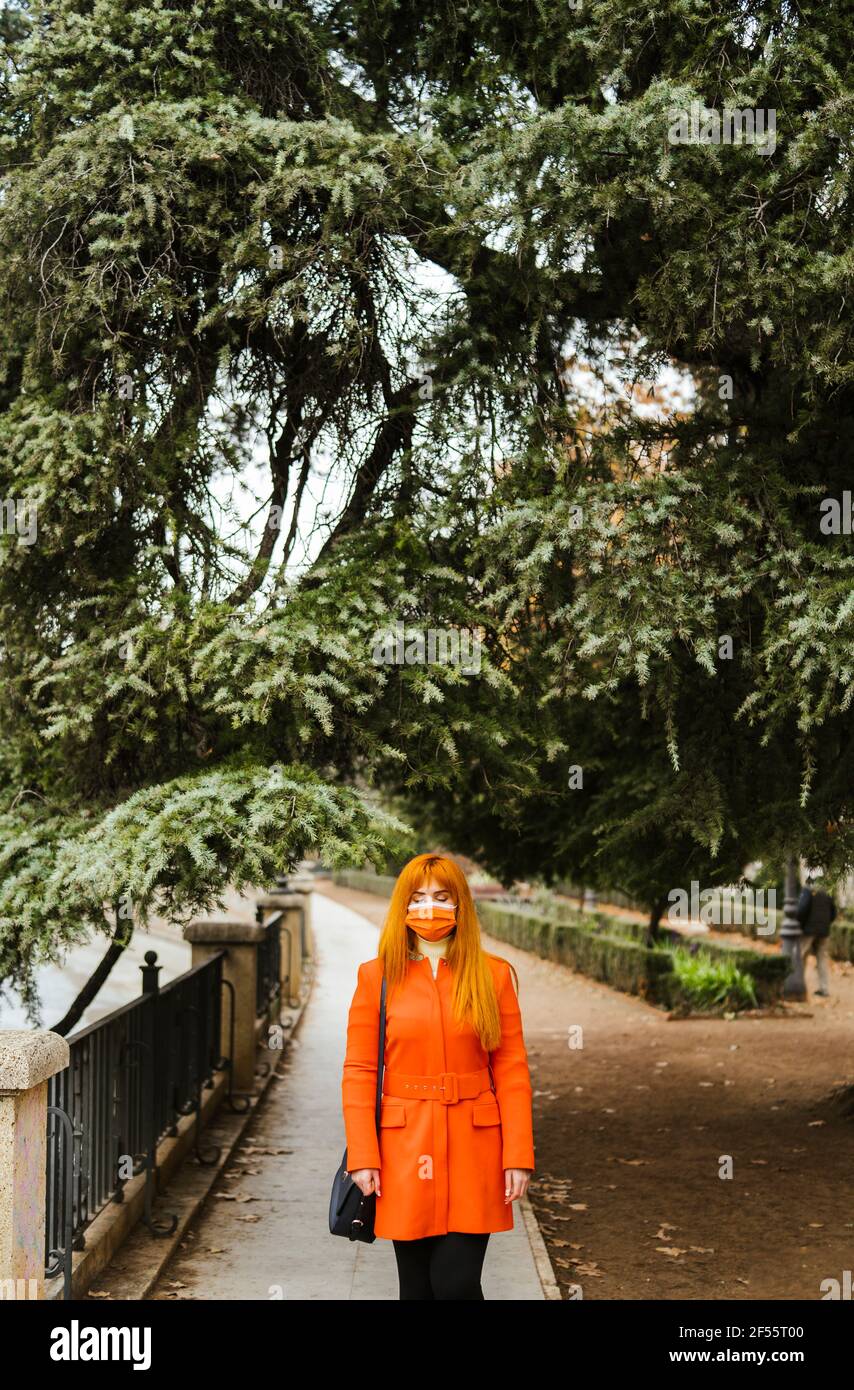Donna con maschera protettiva in piedi contro l'albero al pubblico parcheggio Foto Stock