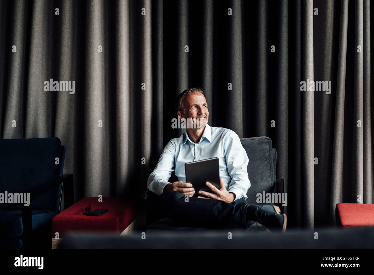 Uomo d'affari sorridente con gambe incrociate al ginocchio che tiene il tablet digitale su poltrona in caffetteria d'ufficio Foto Stock