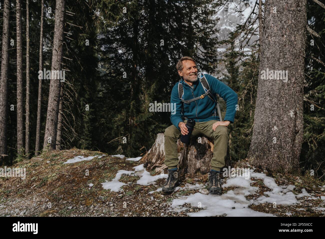 Uomo maturo sorridente che tiene una bottiglia d'acqua mentre si siede contro gli alberi a Salzburger Land, Austria Foto Stock