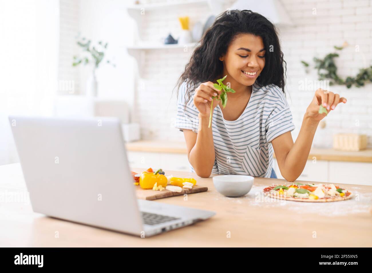 Una ragazza multietnica sorridente carina con capelli ricci impara a cucinare la pizza a casa, a decorarla con i verdi, a preparare cibi sani, a prendersi cura della salute, a mangiare un sacco di vitamine, a sognare di diventare uno chef Foto Stock