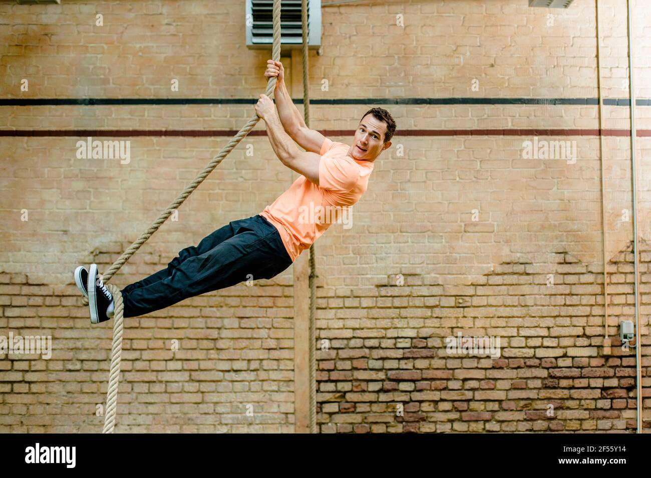 Uomo di sesso maschile appeso sulla corda da muro di mattoni in palestra Foto Stock