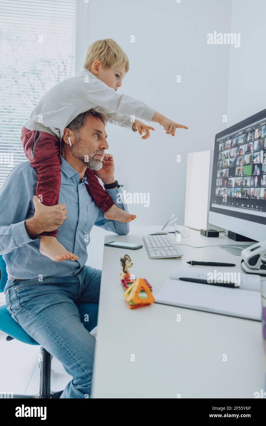 Padre che partecipa alla videoconferenza mentre il figlio è seduto sulla spalla puntare sullo schermo del computer dell'ufficio domestico Foto Stock