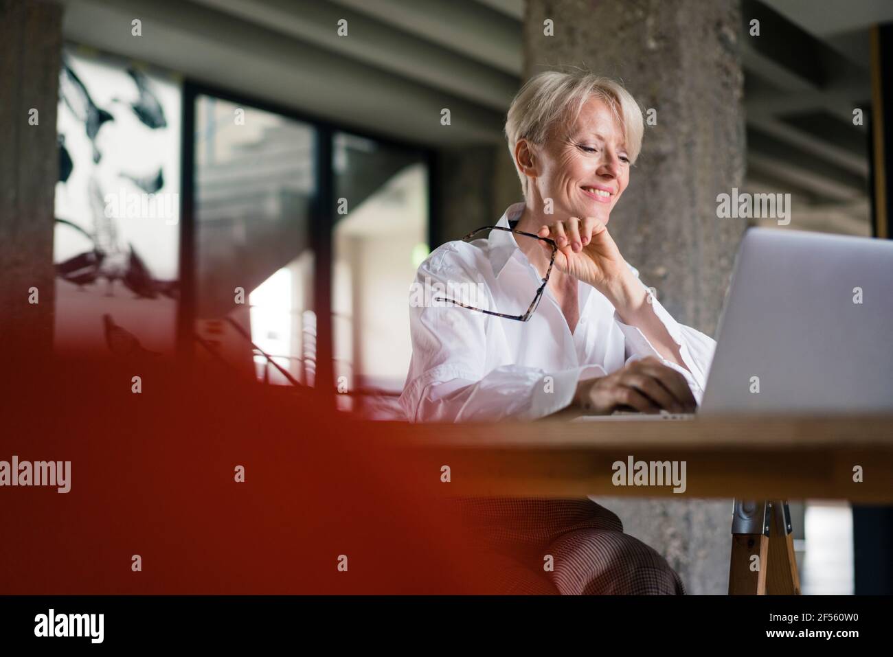 Sorridente lavoratore freelance che tiene gli occhiali mentre usa il computer portatile alla scrivania in ufficio a casa Foto Stock