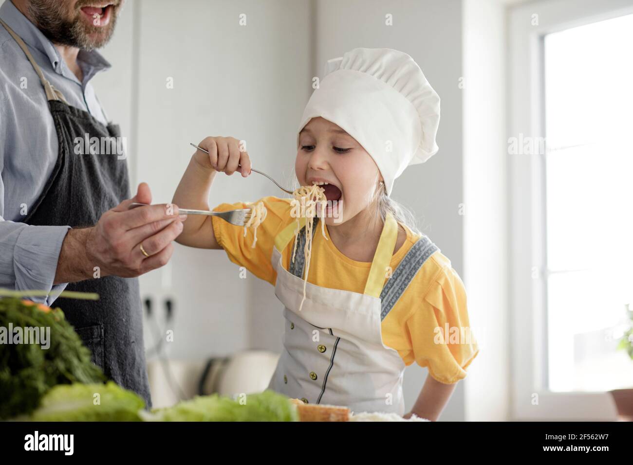 Ragazza che indossa grembiule e cappello dello chef mangiando spaghetti con  il padre mentre si trova in cucina a casa Foto stock - Alamy