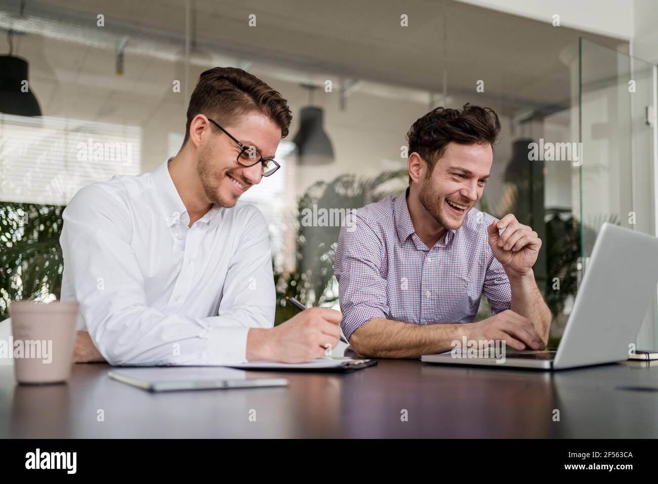 Un uomo d'affari sorridente che lavora un computer portatile mentre un collega maschio scrive sugli appunti in ufficio Foto Stock