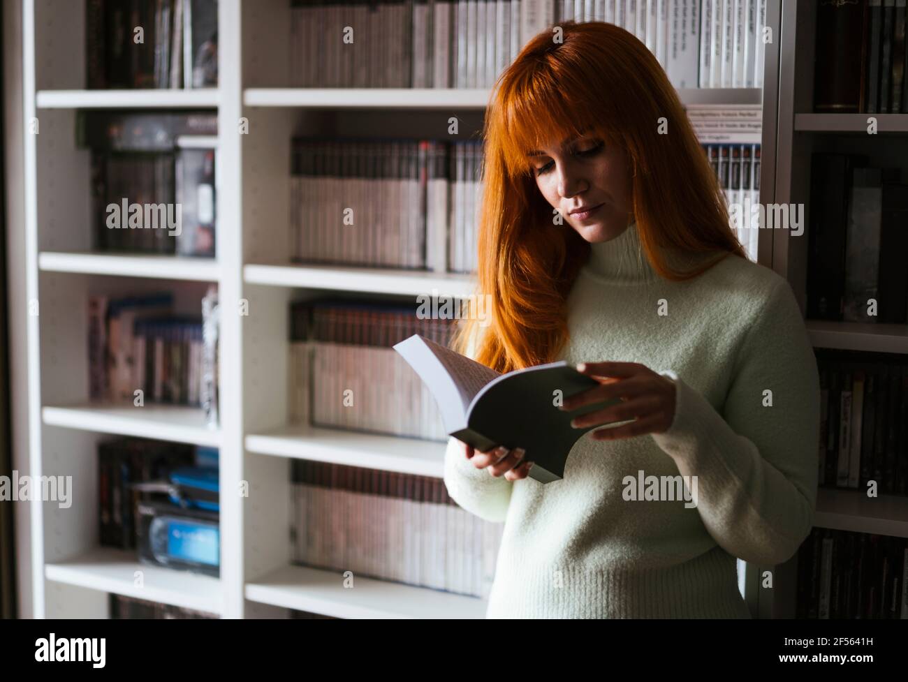 Concentra donna leggere libro contro scaffale alla biblioteca Foto Stock