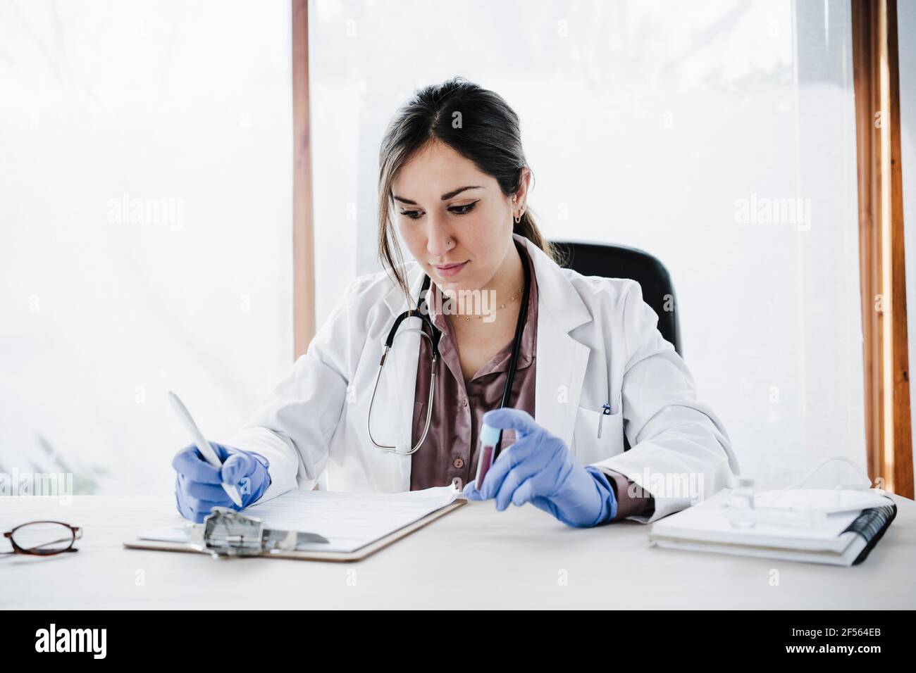 Esperienza medica femminile che scrive i rapporti degli esami del sangue mentre tiene il sangue esempio alla scrivania Foto Stock