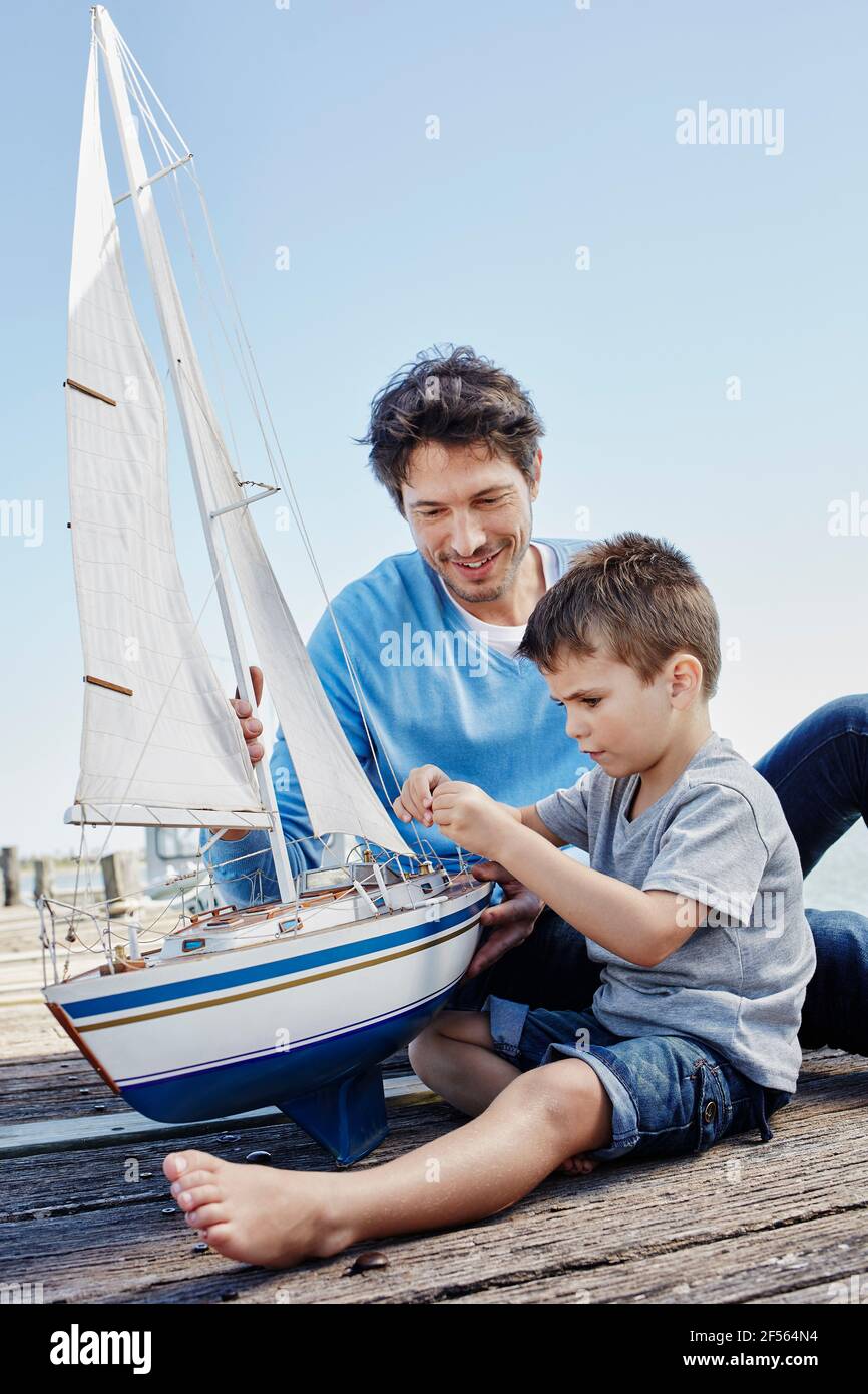 Ragazzo riparazione giocattolo barca a vela mentre si siede da padre sul molo Foto Stock