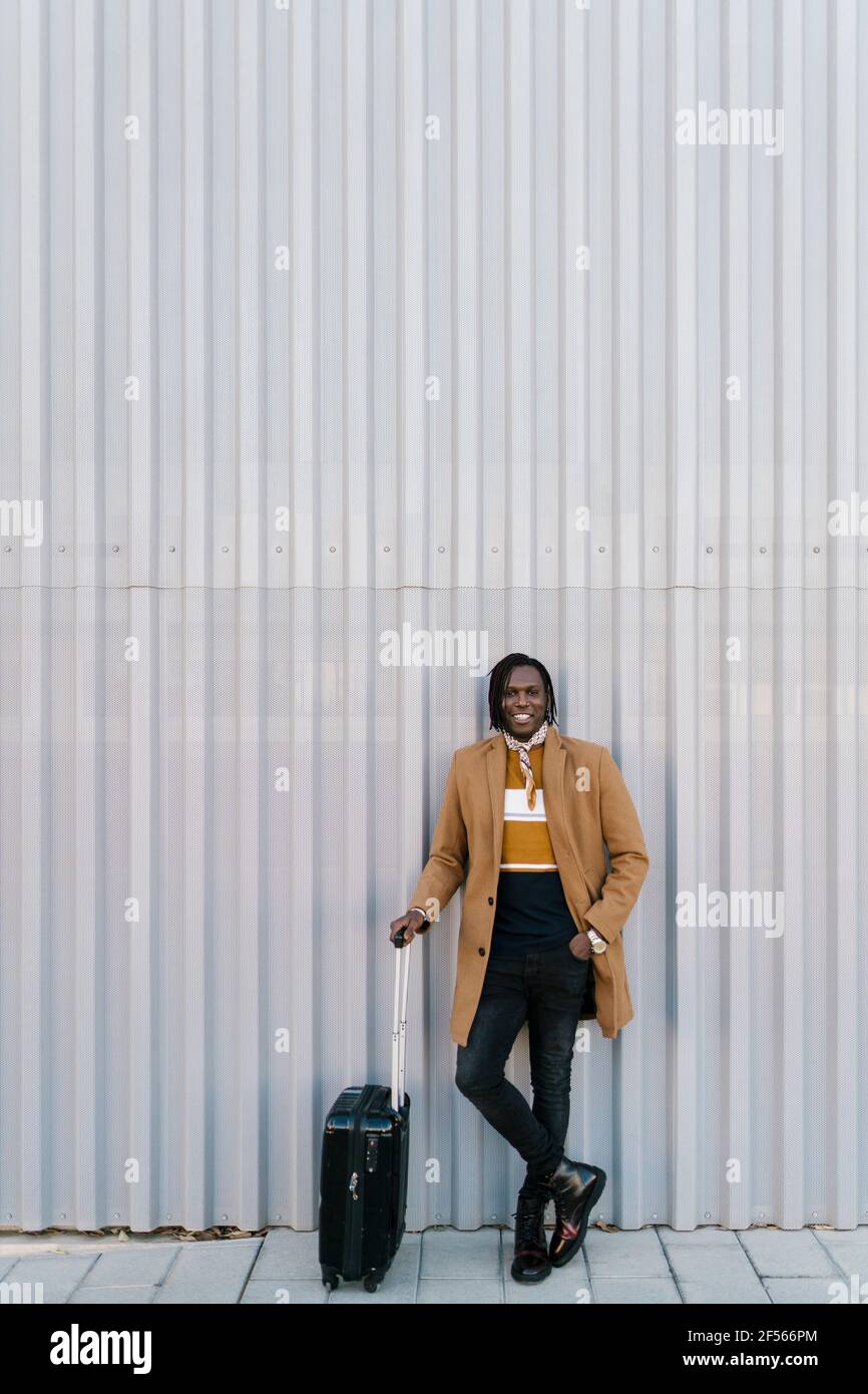 Uomo africano con valigia in piedi mentre si appoggia contro parete grigia Foto Stock