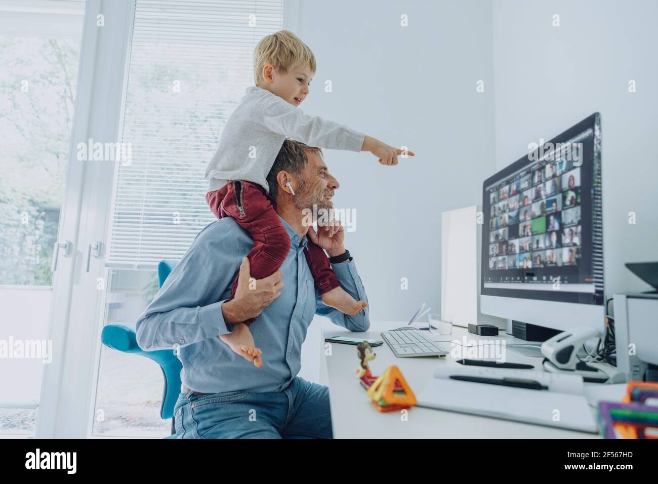 Uomo d'affari maturo che partecipa a una videoconferenza mentre il figlio è seduto sul suo spalla rivolta sullo schermo del computer dell'ufficio domestico Foto Stock
