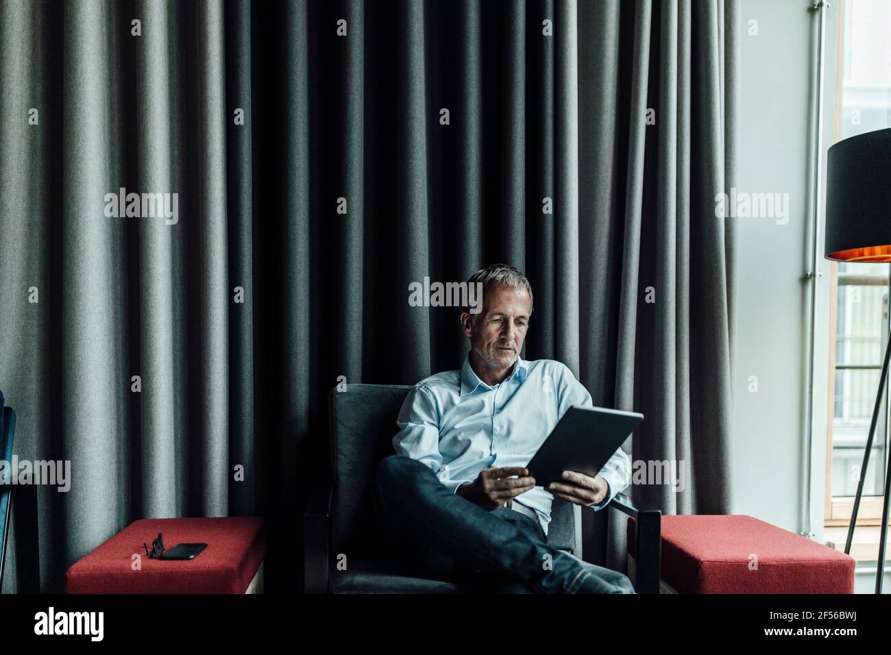 Uomo d'affari serio che usa il tablet digitale contro la tenda grigia in ufficio caffetteria Foto Stock