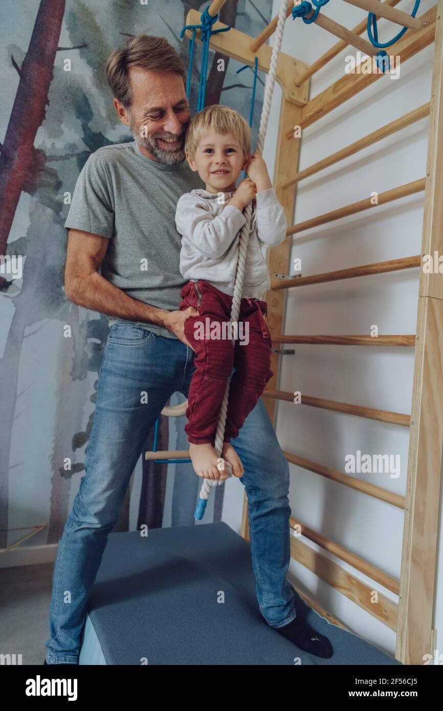 Felice padre che assiste il figlio scalando corda in soggiorno Foto Stock