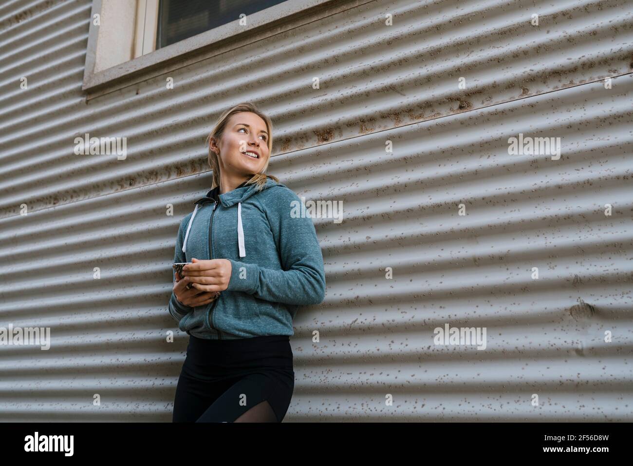 Donna sportiva sorridente con telefono cellulare che guarda lontano mentre si appoggia parete Foto Stock