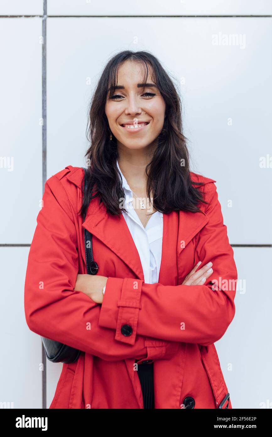 Giovane donna sorridente in trench rosso in piedi con le braccia barrato contro parete bianca Foto Stock