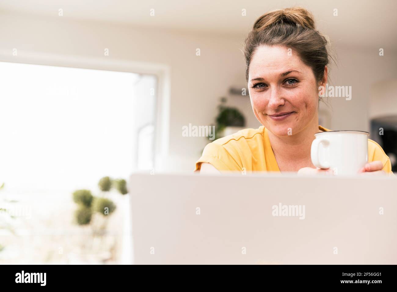 Donna d'affari sorridente che tiene la tazza del caffè mentre prende la pausa dal lavoro a casa Foto Stock