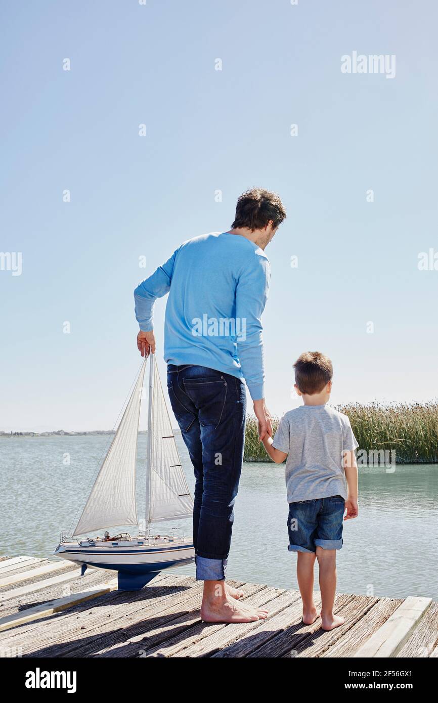 Padre con la barca a vela giocattolo che tiene la mano del figlio mentre si è in piedi sul molo Foto Stock