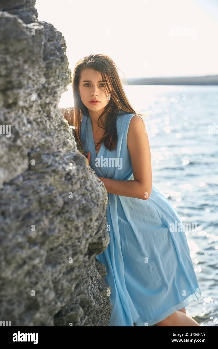 Splendida donna con capelli marroni in piedi vicino alla roccia sul lungofiume. Clima caldo e soleggiato. Concetto di tenerezza e calma. Foto Stock