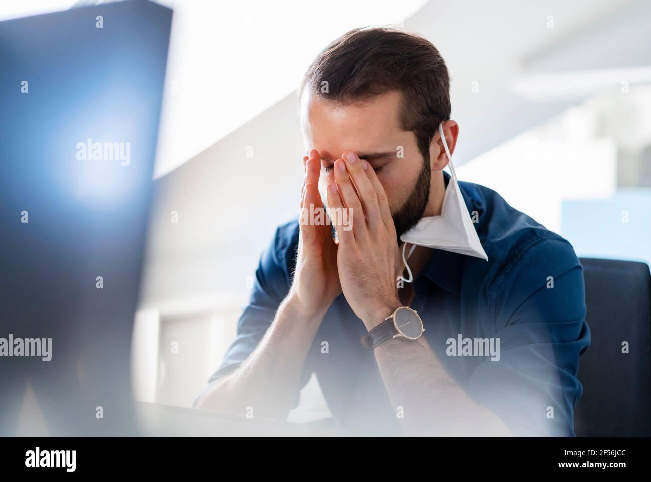 Uomo d'affari con testa in mano e maschera facciale sull'orecchio seduto in ufficio Foto Stock