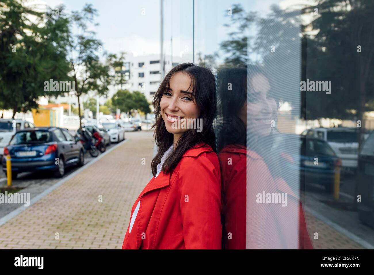 Giovane donna sorridente in trench rosso appoggiato sul vetro finestra Foto Stock