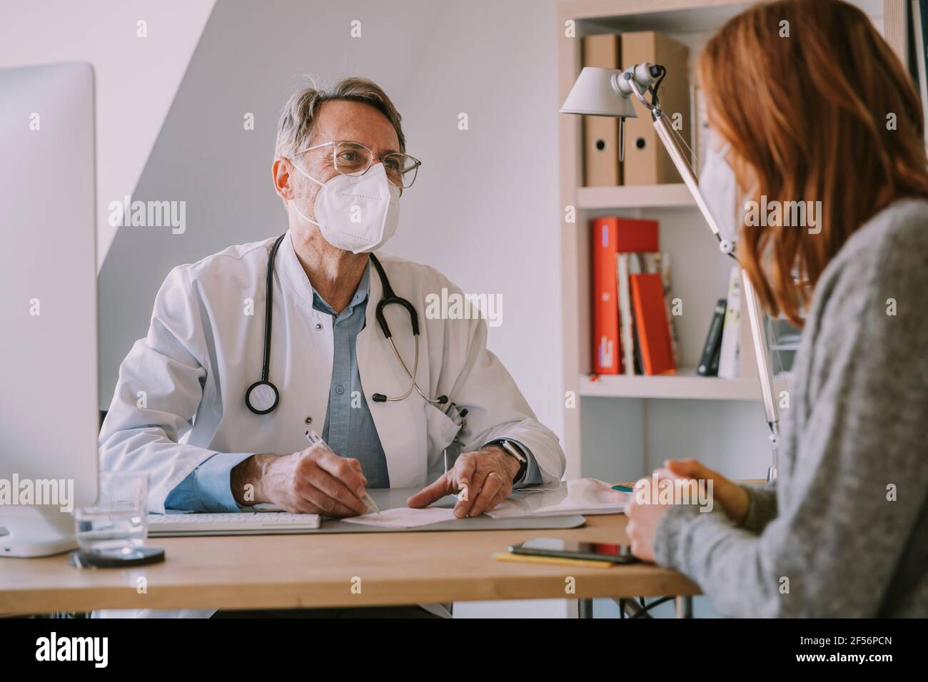 Medico generico maschile con maschera facciale che guarda il paziente mentre seduto all'ufficio del medico Foto Stock