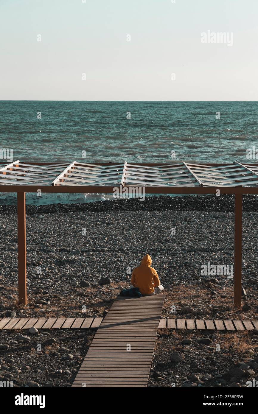 L'uomo Hooded seduto da solo alla fine del lungomare che termina a. bordo di spiaggia costiera rocciosa Foto Stock