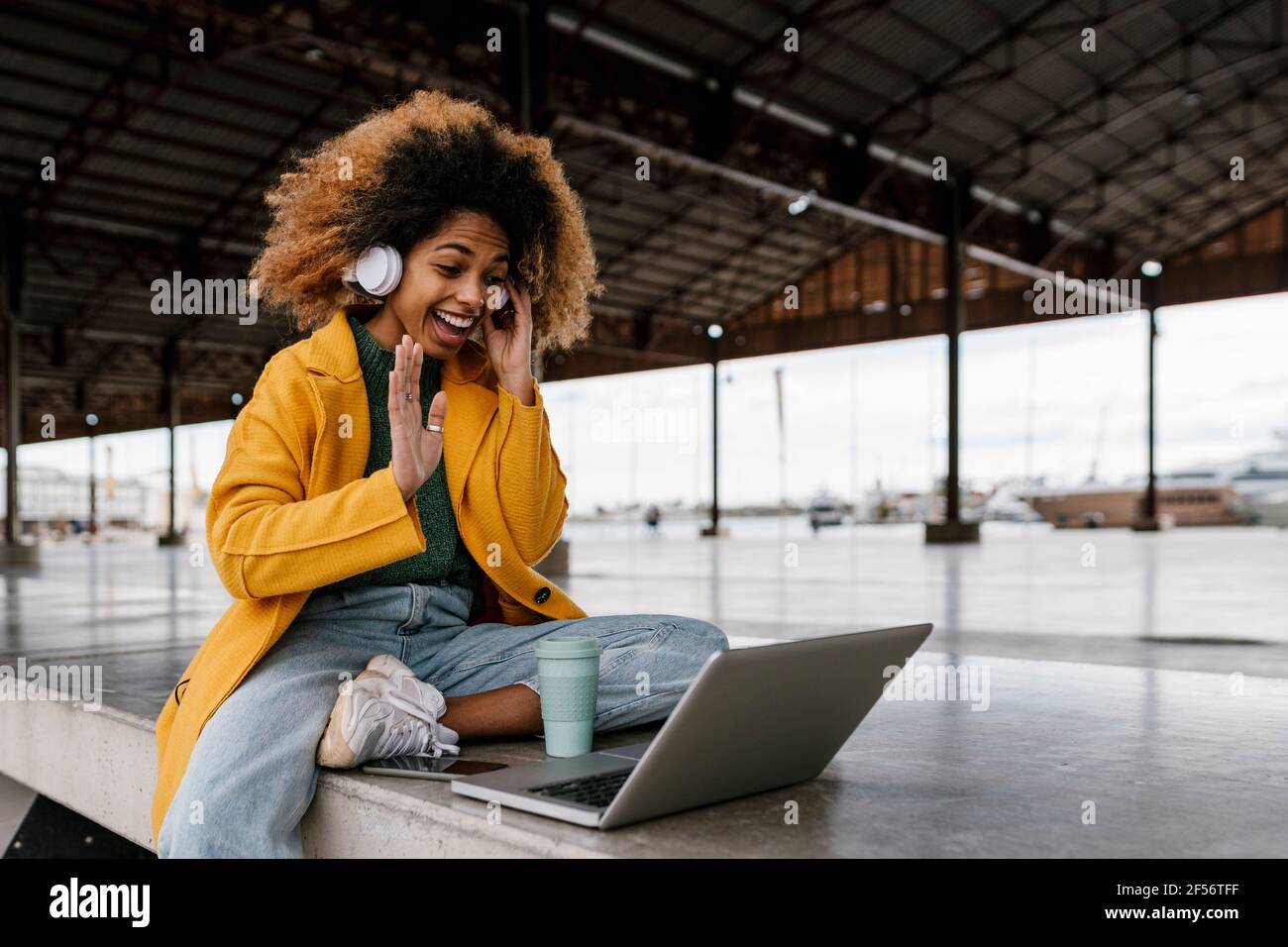 Allegra donna afro con cuffie intrauricolari che ondolano la mano durante il video chiama il computer portatile nell'area salotto Foto Stock
