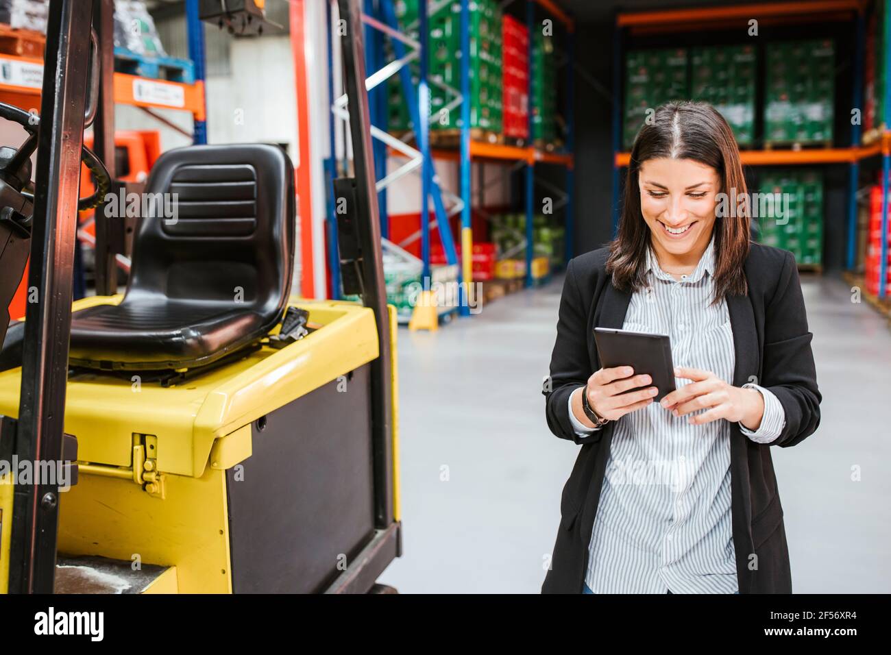 Donna d'affari sorridente che usa un tablet digitale da un carrello elevatore a forche in un magazzino di distribuzione Foto Stock