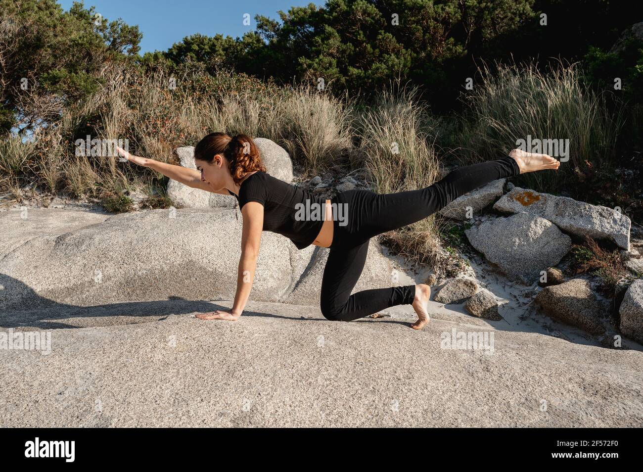 La giovane donna fa esercizi di stretching su una roccia con vegetazione selvaggia sullo sfondo della spiaggia. Sport all'aperto. Foto Stock