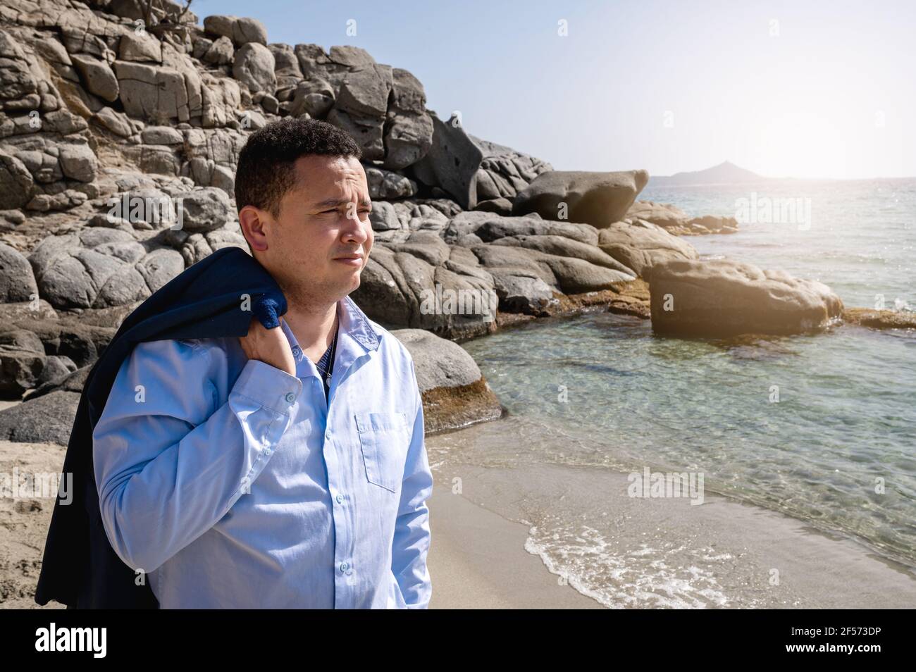 Uomo latino con elegante giacca da decollo tuta camminando in spiaggia sabbiosa e guardando lontano. Fuggi dalla città per trovare un luogo tranquillo. Foto Stock