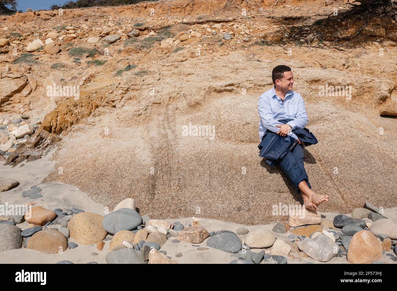 Latino elegante uomo d'affari seduto su una roccia alla spiaggia guardando l'orizzonte al suo fianco. Relax alla ricerca di un luogo tranquillo fuori dal caos del c. Foto Stock