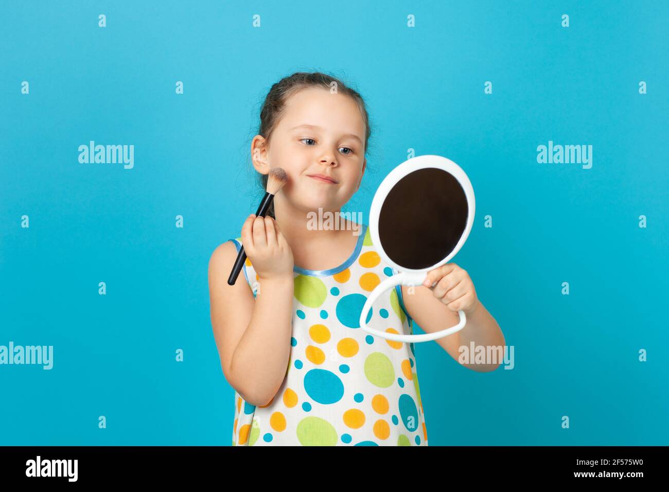 primo piano ritratto di una ragazza sorridente che talcola le guance con un grande pennello e tiene in mano uno specchio bianco, isolato su uno sfondo blu Foto Stock