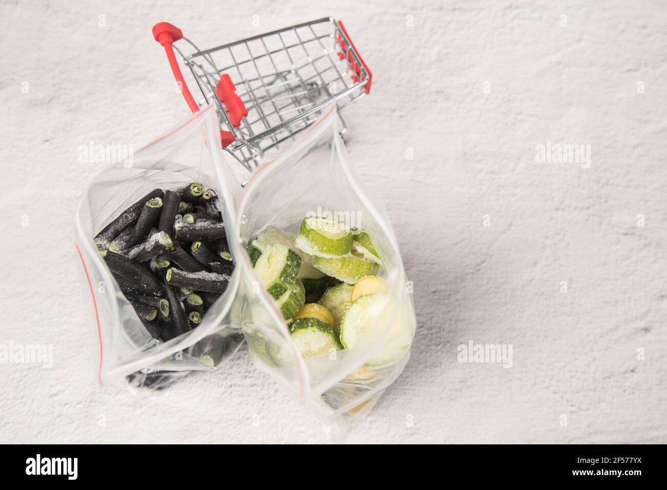 Sacchetti di zucchine surgelate e fagioli a corda si trovano su uno sfondo chiaro accanto a un mini carrello del supermercato. Foto Stock