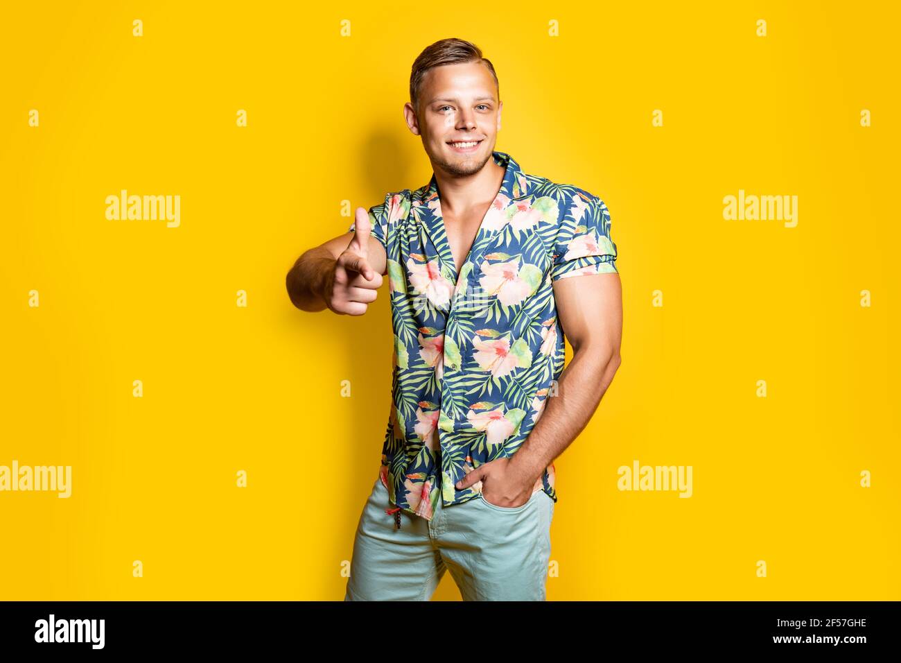 Positivo giovane maschio nel quartiere alla moda di outfit sorridente e puntando alla telecamera mentre in piedi contro sfondo giallo Foto Stock