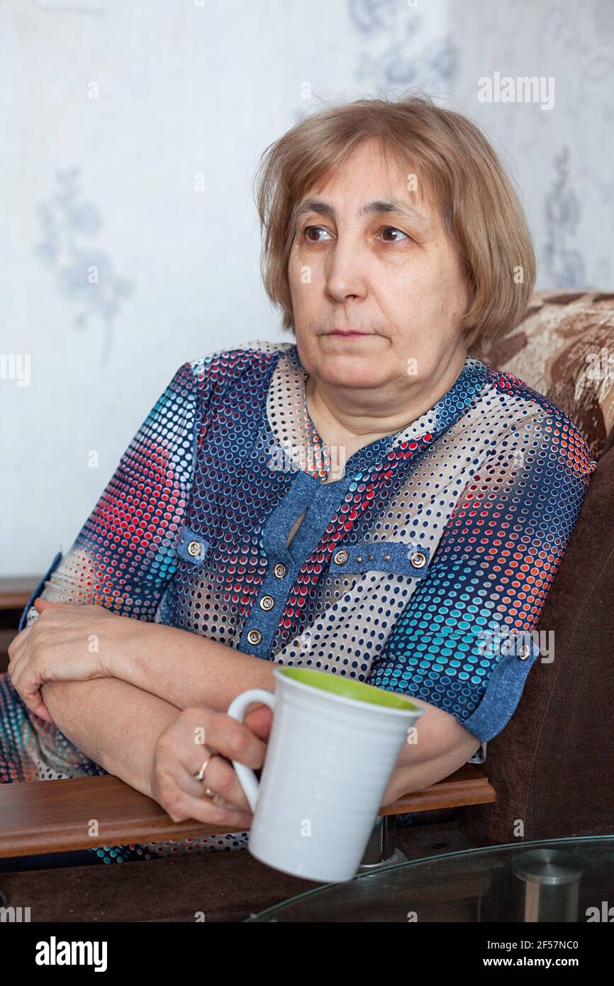 Donna caucasica matura che tiene una tazza da tè, seduta in poltrona in una stanza domestica Foto Stock